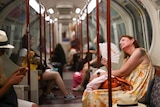 A woman looking very hot and sweaty fans herself with a paper fan while sitting on the London tube