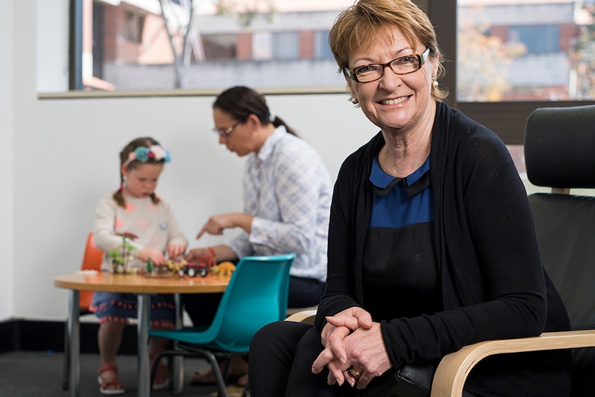 A woman sitting on a chair with a woman and child in the background.