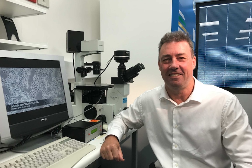 A man sitting next to a microscope and a screen showing cells.