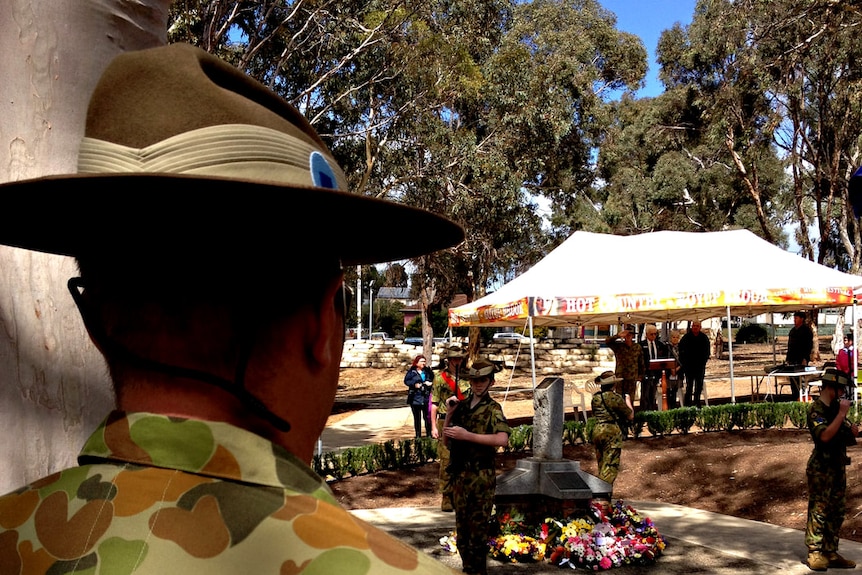 A army person standing with his back to the camera in the foreground with younger army corps standing around a memorial outside