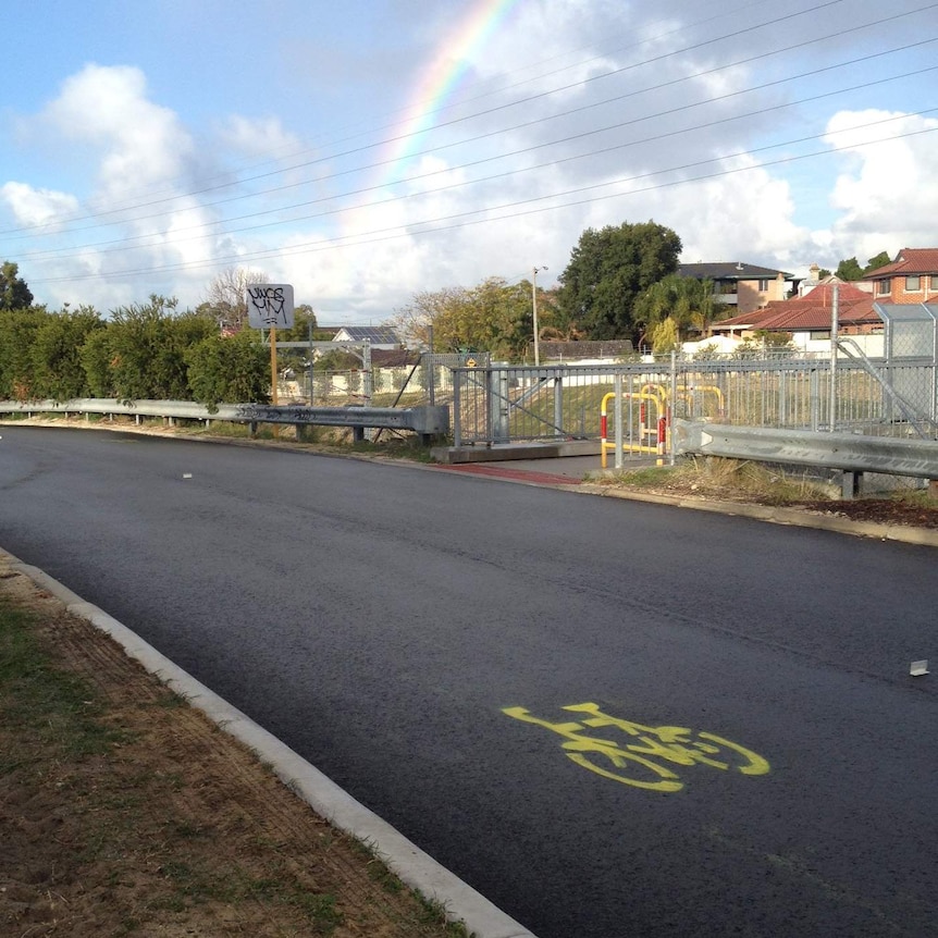 A yellow bike priority marking spray painted on Rutland Avenue in Victoria Park in Perth with a rainbow in the sky above.