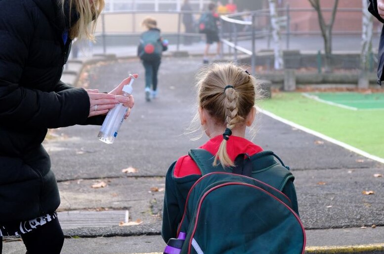 A woman stands at the school gate and offers hand sanitiser to a primary school student.