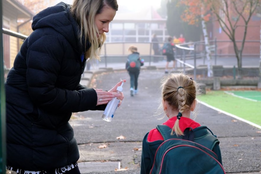 A woman stands at the school gate and offers hand sanitiser to a primary school student.