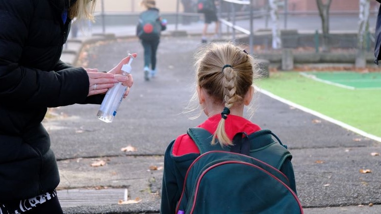 A woman stands at the school gate and offers hand sanitiser to a primary school student.