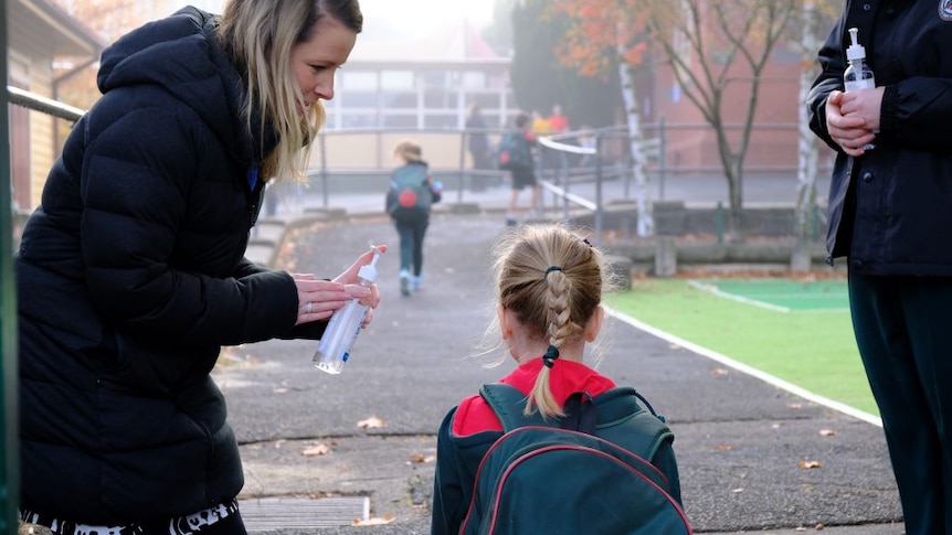 A woman stands at the school gate and offers hand sanitiser to a primary school student.