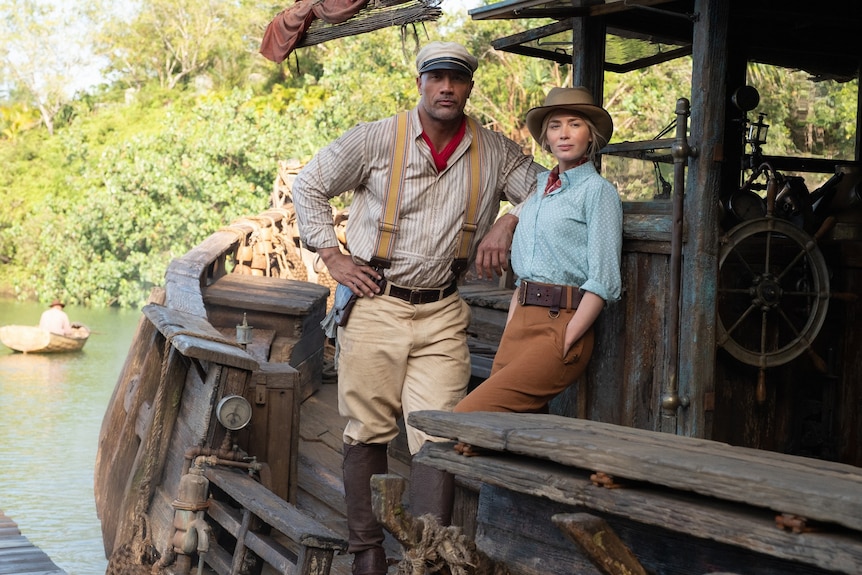A confident-seeming Dwayne Johnson and Emily Blunt lean against the side of the helm of a wooden boat floating on a river