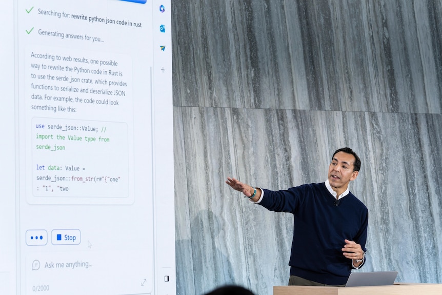 A middle-aged man stands at a lecturn, gesturing with one arm to a projector screen displaying Microsoft's Bing AI chatbot.