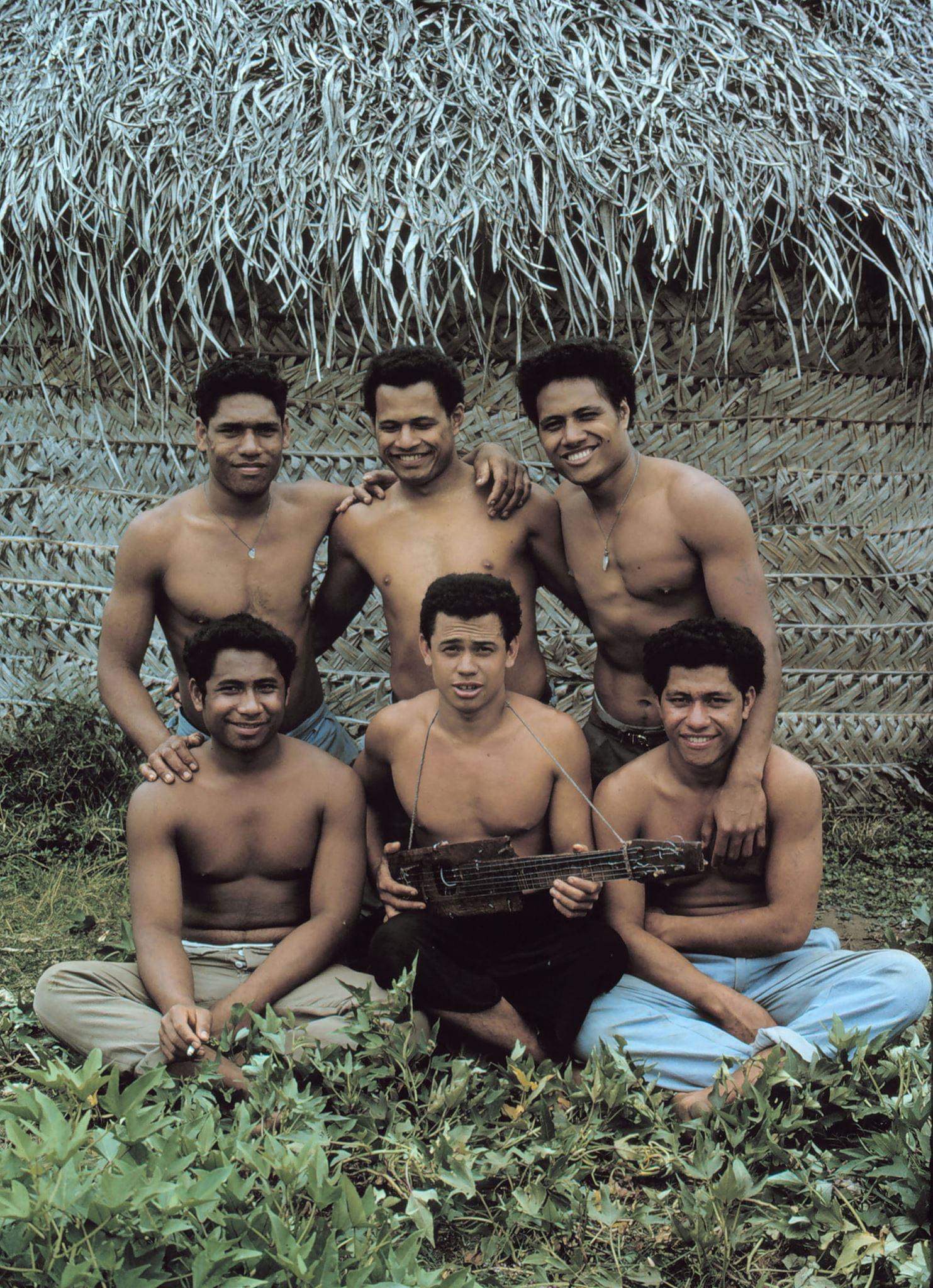 Six, smiling, well-built shirtless men in front of a thatched roof hut.
