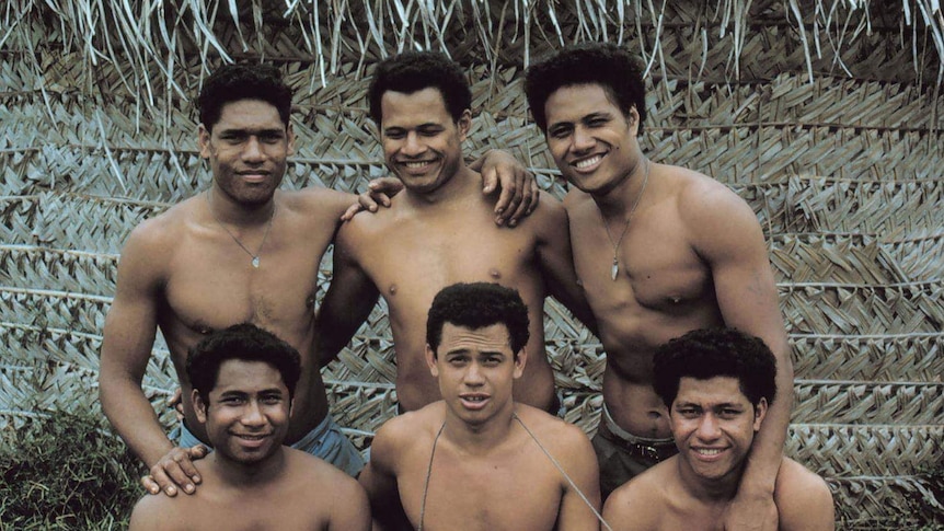 Six, smiling, well-built shirtless men in front of a thatched roof hut.