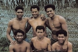 Six, smiling, well-built shirtless men in front of a thatched roof hut.
