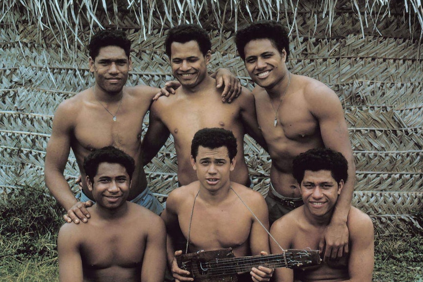 Six, smiling, well-built shirtless men in front of a thatched roof hut.