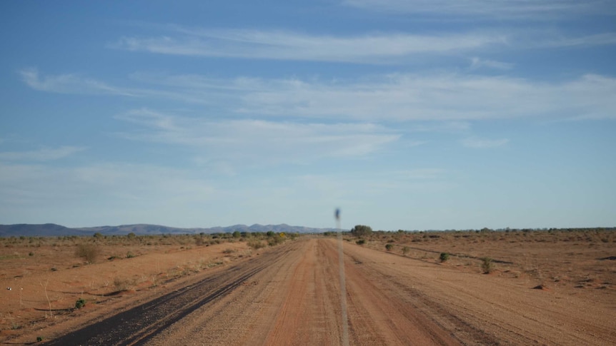 A dusty outback road, seen through the windscreen of a car with UHF aerial and bonnet in the foreground.