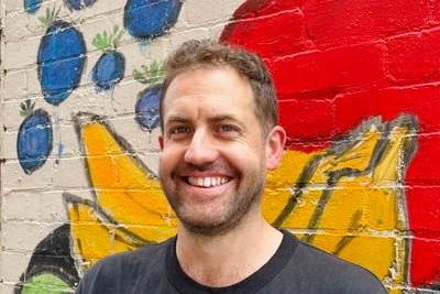 A man wearing a t-shirt standing in front of a mural on a brick wall smiling