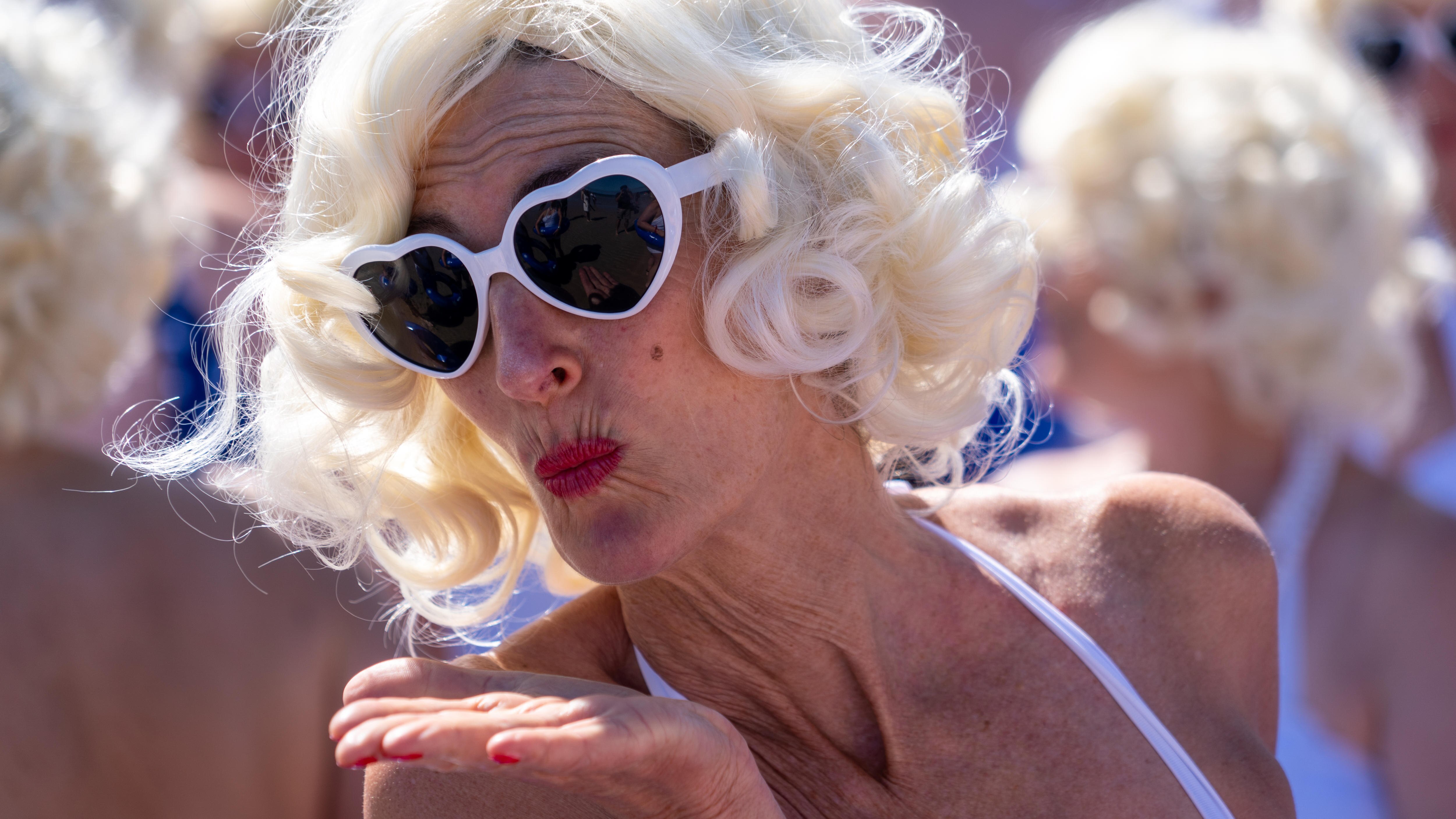 A person wearing a blonde curly wig, white heart-shaped sunglasses and white swimwear blows a kiss to the camera