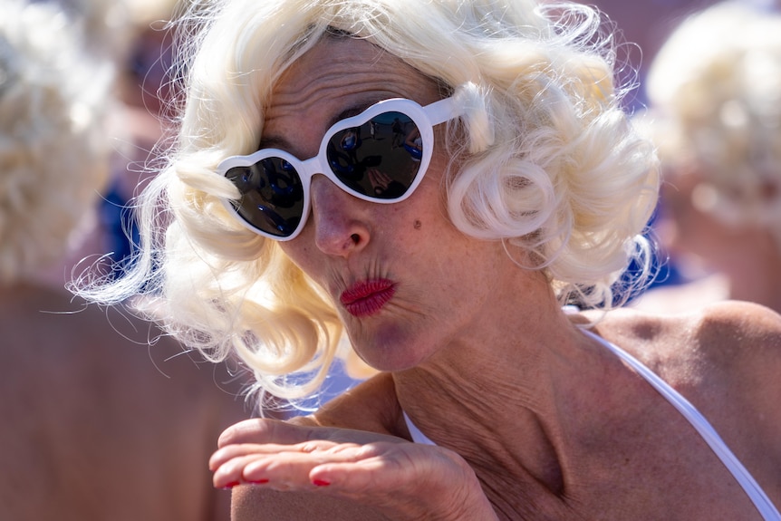 A person wearing a blonde curly wig, white heart-shaped sunglasses and white swimwear blows a kiss to the camera