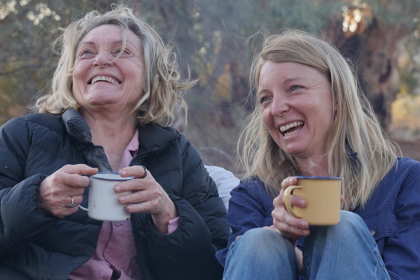 A mid-shot of two women sitting at a campsite drinking from camping mugs looking off-camera and laughing.