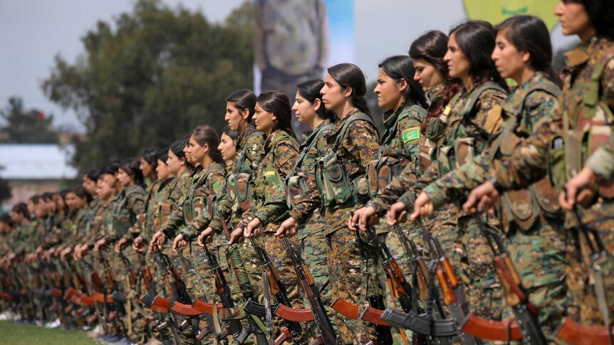 Kurdish female fighters stand in a line holding guns at their sides.