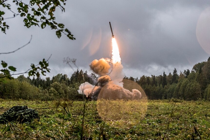 A Russian Iskander-K missile takes off in a field surrounded by trees.