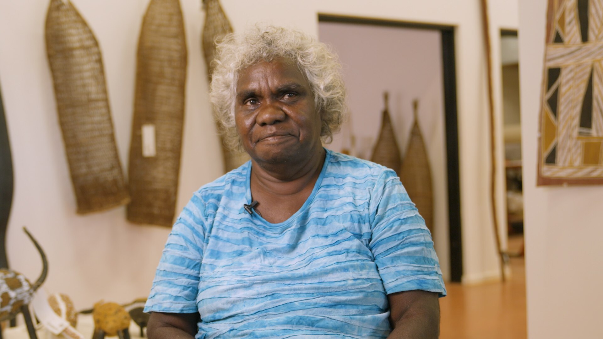 Doreen Jinggabbarra, a Burarra woman in her 60s with short curly grey hair, wearing a bright blue shirt in a gallery.
