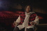 Middle-aged black woman with short corn rows wears a thick beige jumper underneath a red and black checkered vest in a theatre.