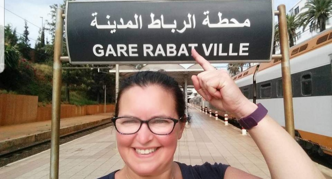 Penny Rabarts on her travels in Morocco, standing below a train station sign reading 'Gare Rabat Ville'.