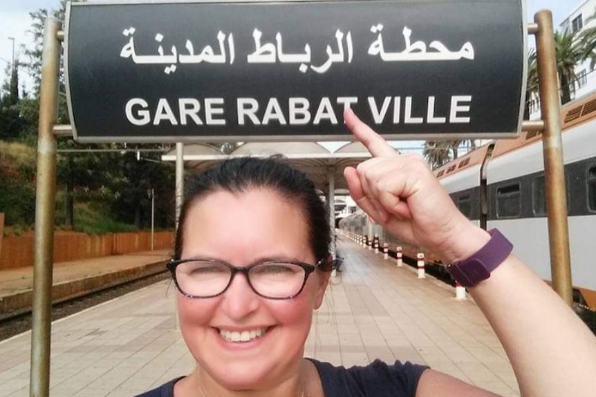 Penny Rabarts on her travels in Morocco, standing below a train station sign reading 'Gare Rabat Ville'.