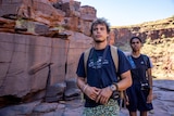 A young man stands in a red canyon in the Australian outback, holding a camera. A young Indigenous woman stands behind him,