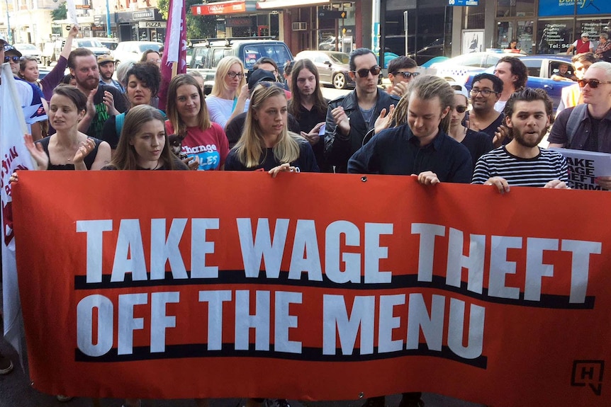 Workers and union members hold up an orange banner that says 'Take wage theft off the menu'.