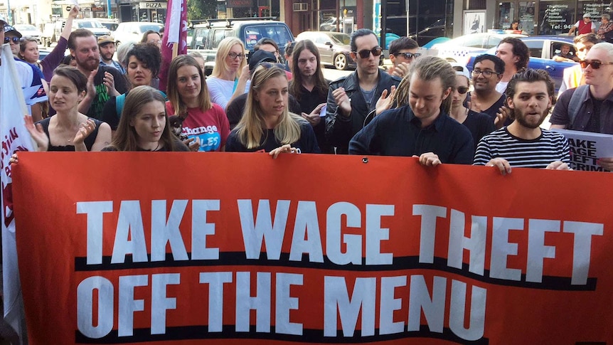 Workers and union members hold up an orange banner that says 'Take wage theft off the menu'.