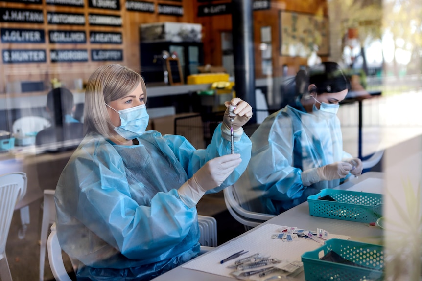 Two women wearing blue PPE hold syringes and vials with a wooden bar visible in background