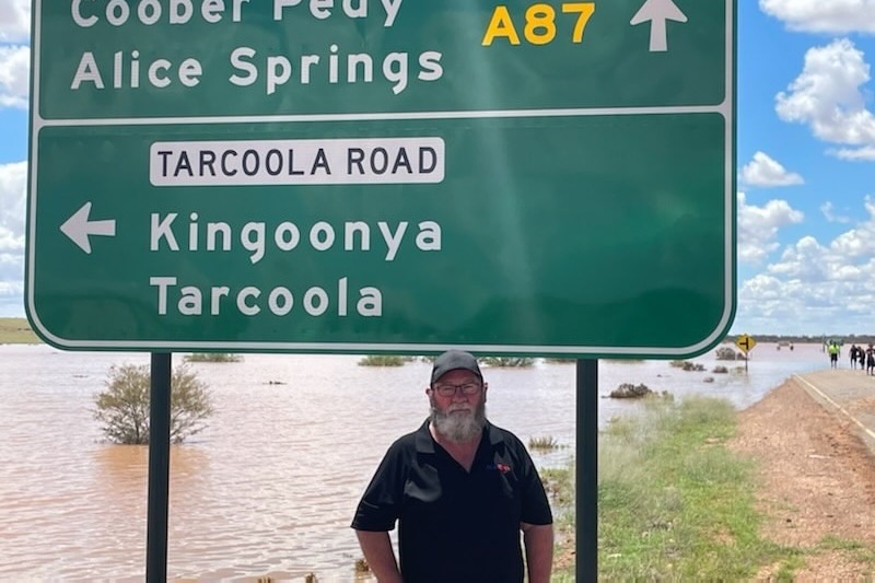 A man with a beard and wearing a cap stands underneath a  road sign, with flooding on the road shoulder behind him.