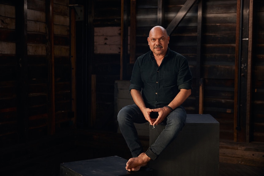 A 50-something Indigenous man in jeans and a button-down sits looking into the camera, resting his hands on his legs