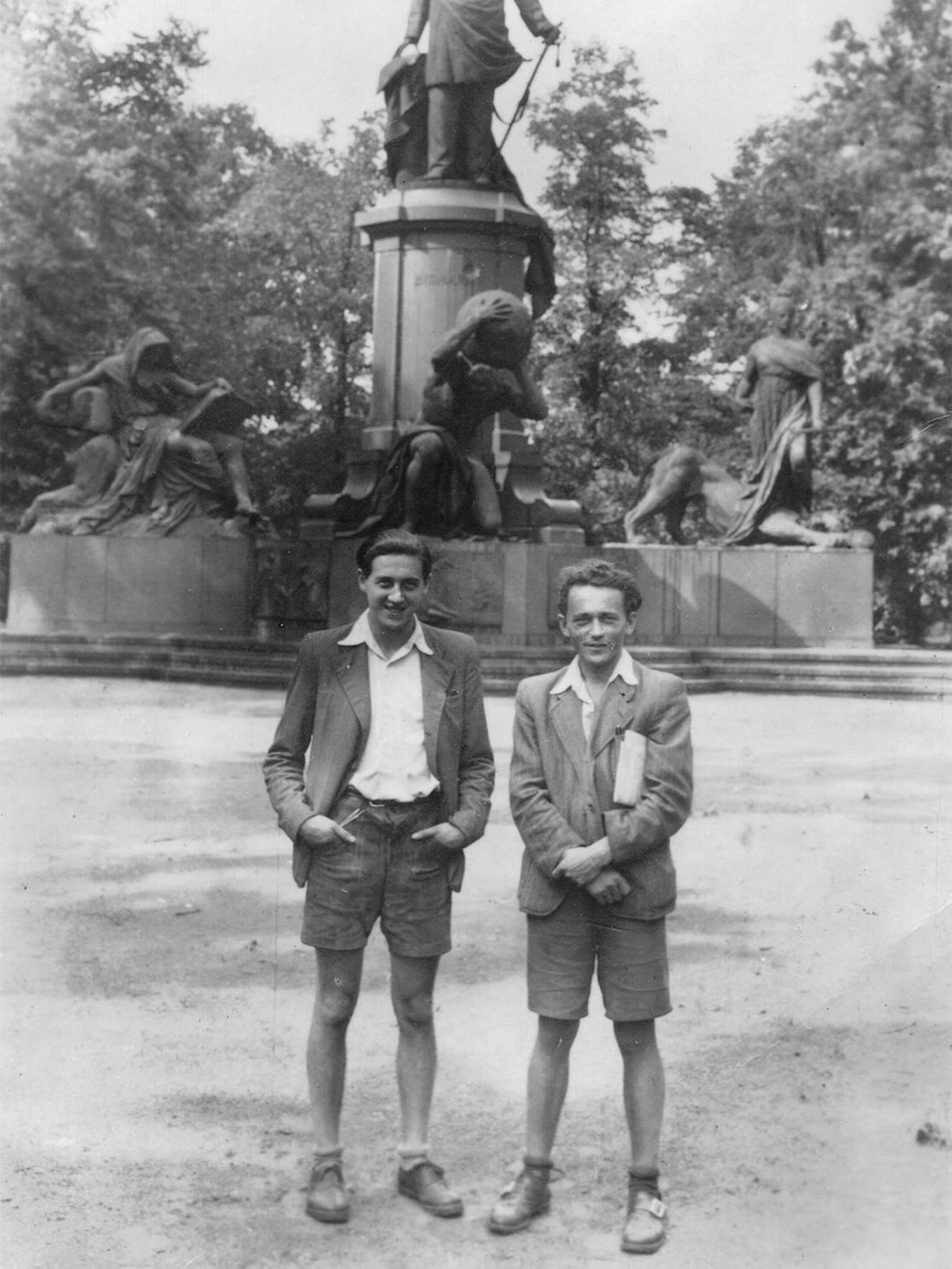 Two young men wearing shorts stand in front of a statue in a park in a black and white photo.