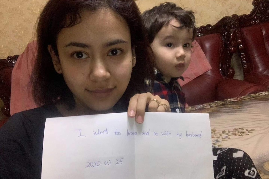 A woman in a selfie with her toddler son. She holds piece of paper reading: "I want to leave and be with my husband. 2020.02.25