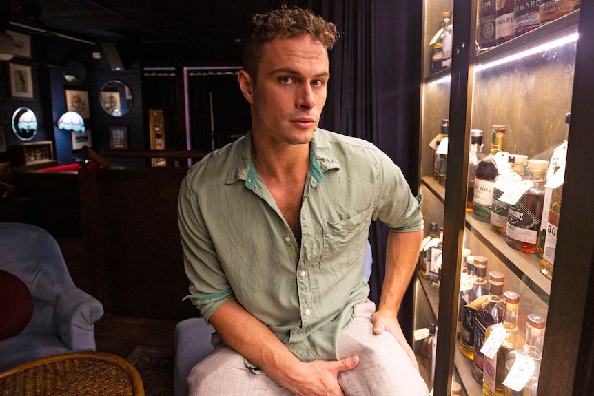 A young man sits in a bar with bottles on the wall, looking straight at the camera. 