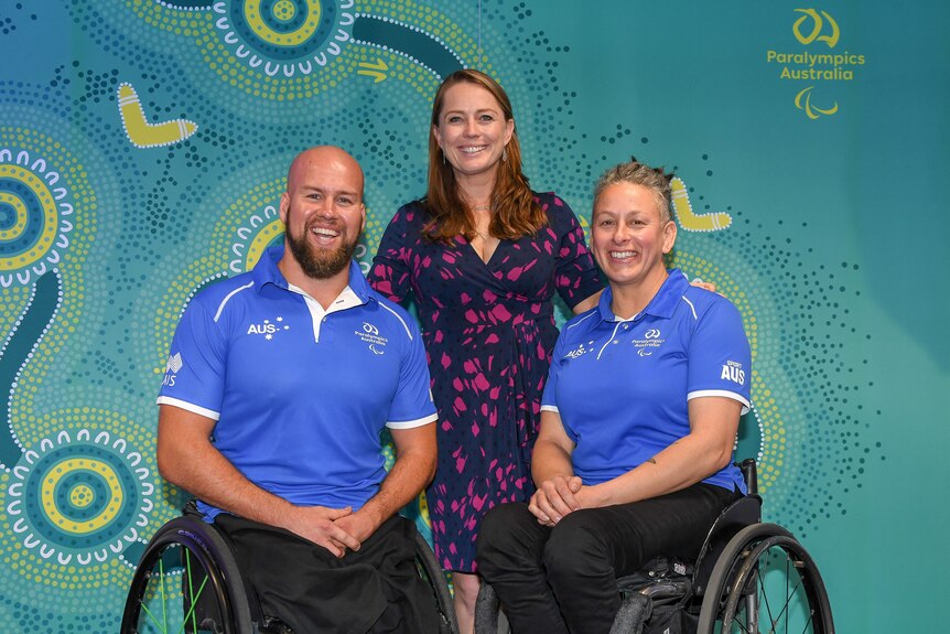 A woman stands between two people in wheelchairs, all are smiling and posing