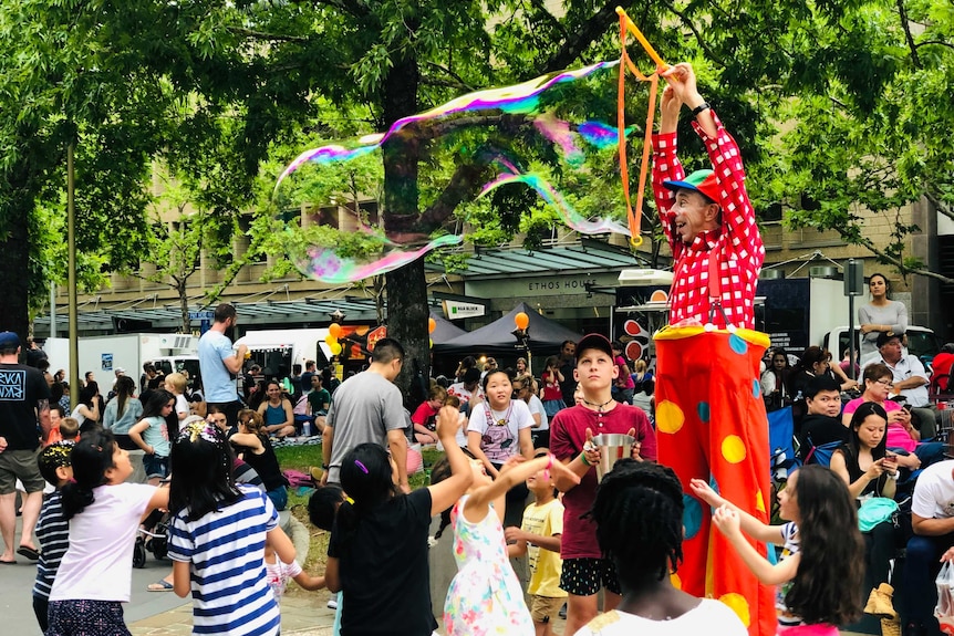 A clown on stilts blows a large bubble while a group of small children look on.