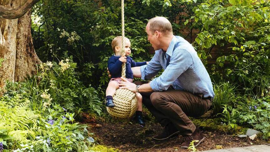 Prince William crouches down to play with his son Prince Louis on a rope swing surrounded by foliage