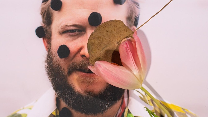 Justin Vernon's bearded face, obscured by a leaf, a flower and pom-poms glued to his head