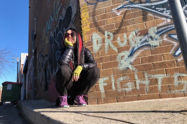 Emma Jane squats next to a wall that has "drugs & fights" written on it.