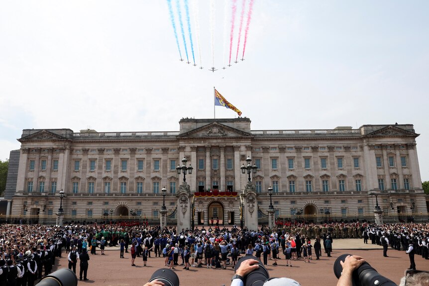The Red Arrows fly over Buckingham Palace with crowd watching.