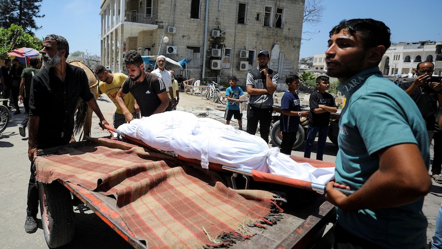 A body wrapped in a white sheet is lifted onto the back of a trailer by two young Palestinian men.