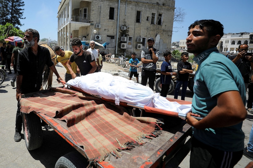 A body wrapped in a white sheet is lifted onto the back of a trailer by two young Palestinian men.