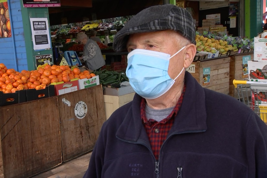 A man wearing a cap and a blue surgical mask stands in front of a fruit and veg shop