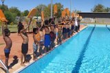 Children stand on the side of a pool, each holding a yellow kickboard.