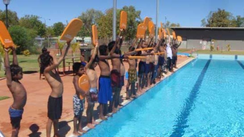 Children stand on the side of a pool, each holding a yellow kickboard.