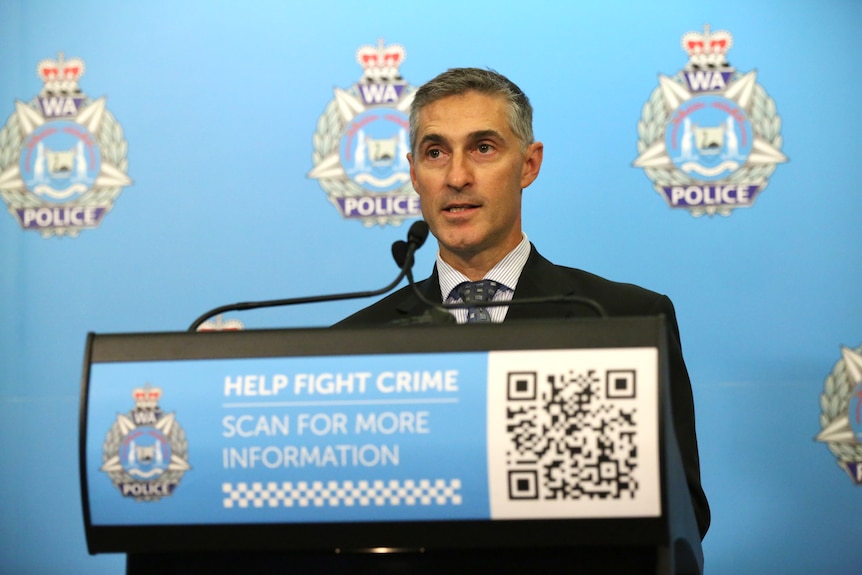 A man in a black suit standing behind a podium. A WA police banner is in the background.