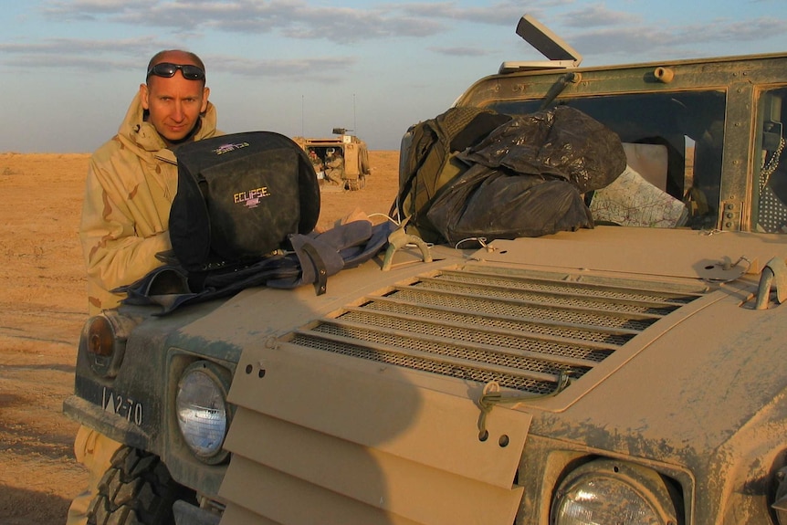 A man in military gear stands near the front of an army vehicle.