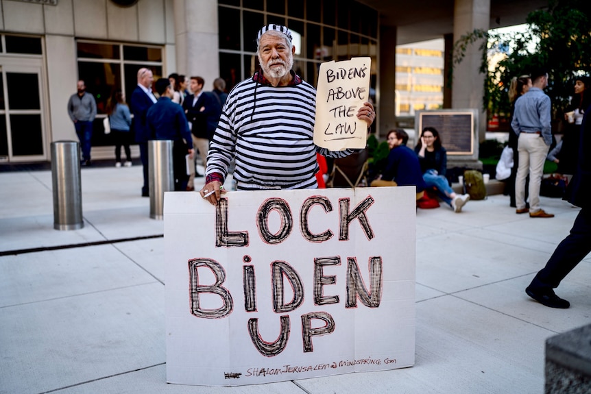 A bearded man wears a black-and-white striped prisoner costume and holds signs with messages including 'Lock Biden Up'