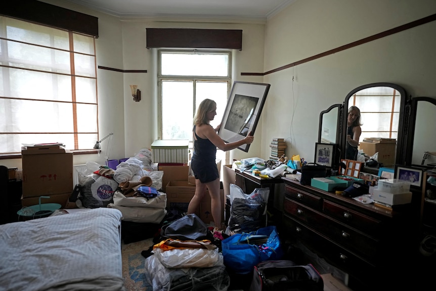 A woman in dark singlet top and shorts moves a large picture frame. There are many packed bags in the room